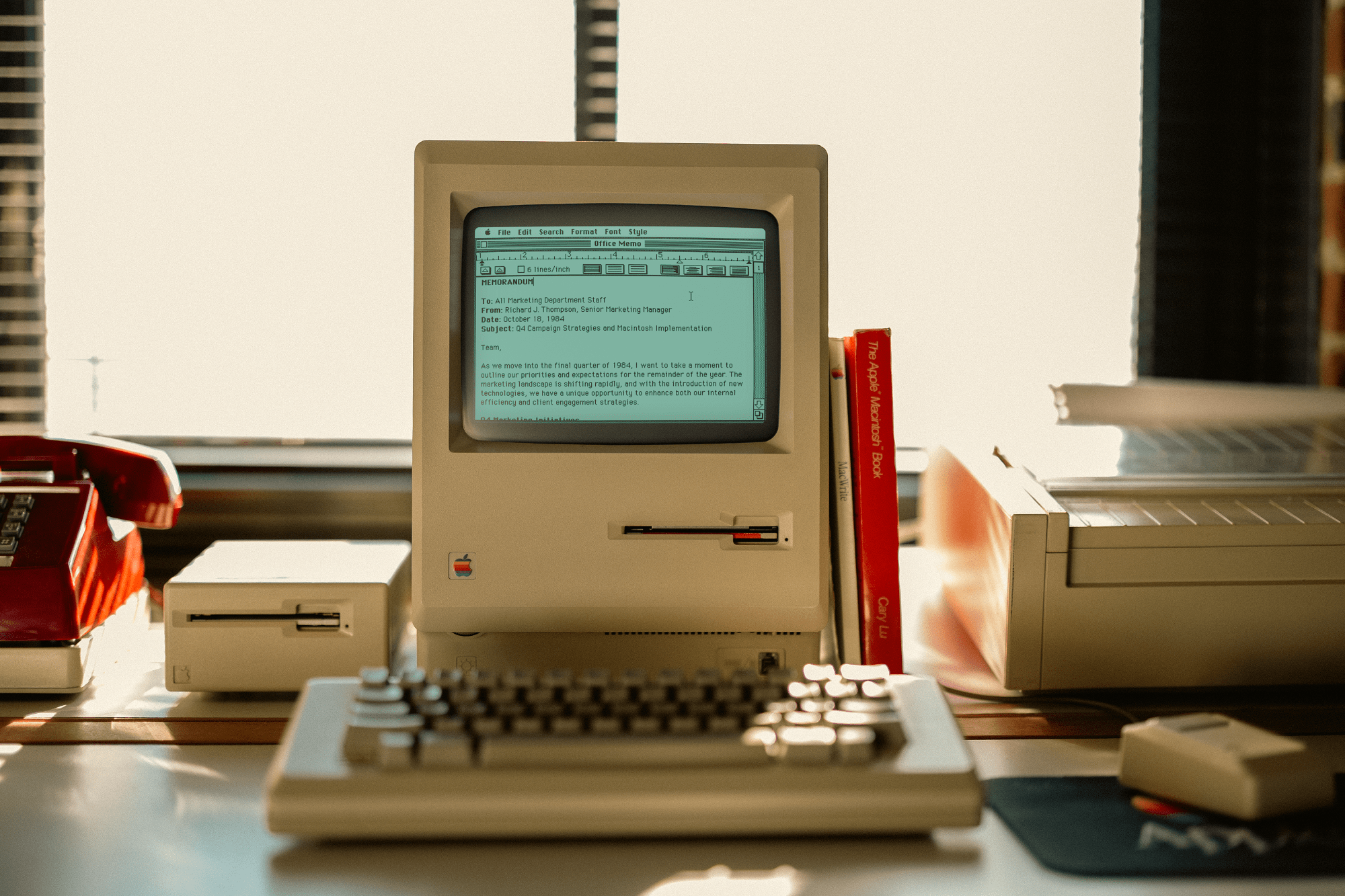 A vintage Apple desk setup with an all-in-one Macintosh (M0001) displaying a 1984 memo, a Macintosh keyboard (M0110), a Macintosh mouse (M0100), a 400K external drive, an ImageWriter printer, and a red rotary phone by the window