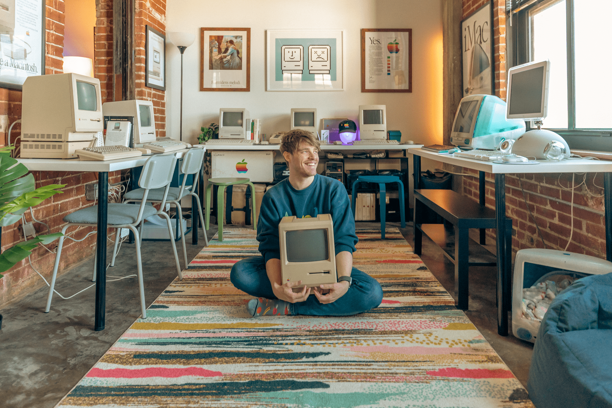 Chris Brueggeman, a video editor based in St. Louis, MO, US, sitting cross-legged on a colourful rug holding an all-in-one Macintosh (M0001) in a room filled with vintage Apple computers, including iMac G3 models and Macintosh Classics