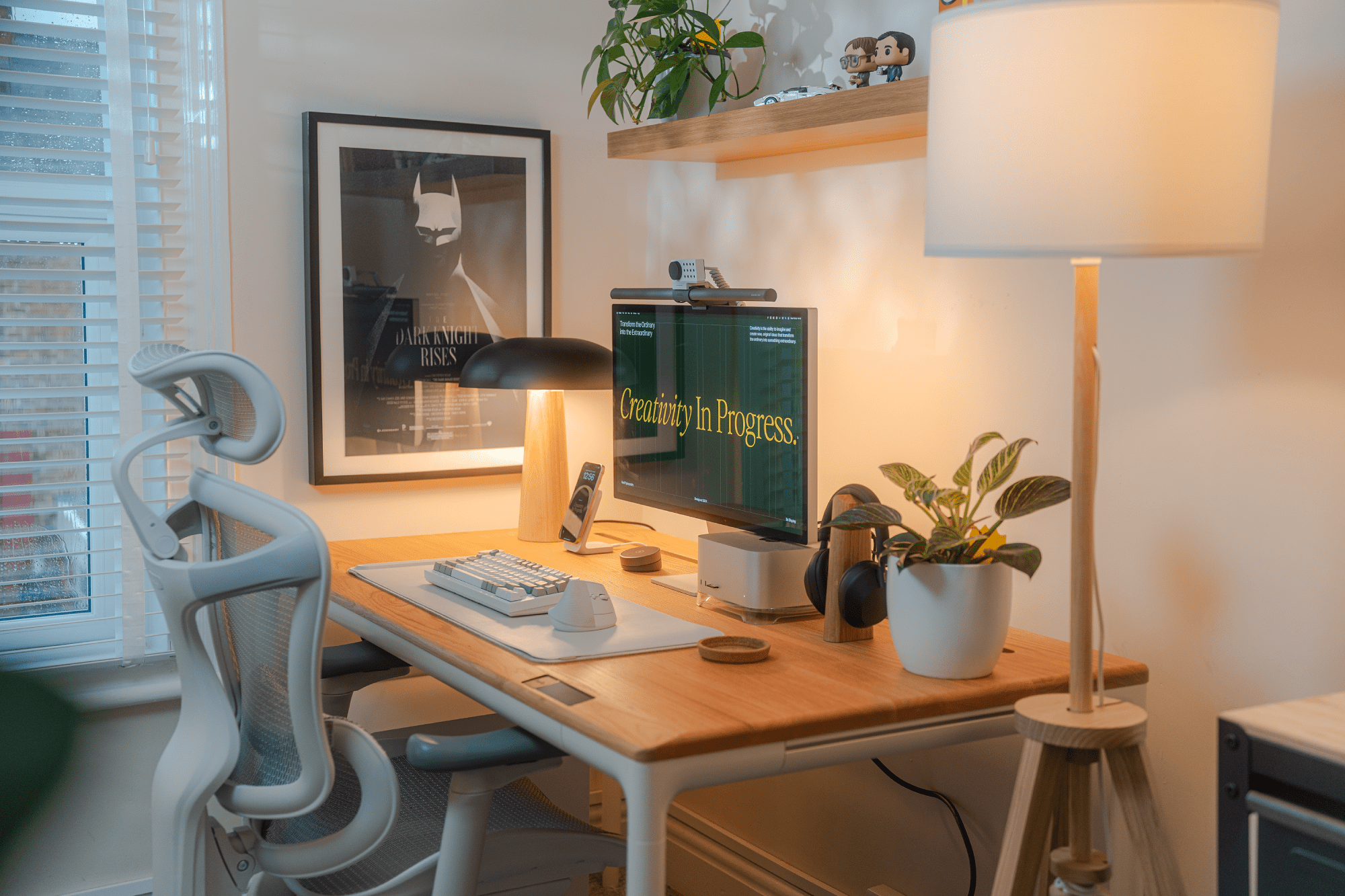 A tidy workspace setup with ergonomic chair, warm lighting, plants, and a framed Batman poster on the wall